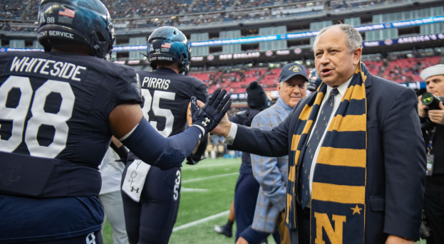 Ein Mann im Anzug und Krawatte gibt einem Fußballspieler die Hand auf einem Feld, umgeben von einer Gruppe von Menschen, mit einer Person, die eine Kamera auf der rechten Seite hält, und einem Stadion mit Menschen und Schildern im Hintergrund, die "Notre Dame Football Coach Mike McCarthy und die Fighting Irish" lesen.