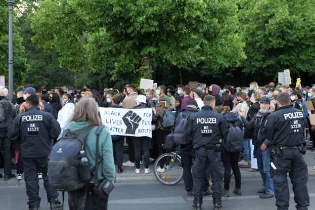 Eine große Gruppe von Menschen steht auf der Straße und hält Schilder und trägt Mützen und Taschen, mit einem Fahrrad im Vordergrund und Bäumen und einem Pfahl im Hintergrund, bei einer Black-Lives-Matter-Demonstration in Berlin.