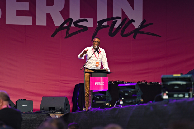 Ein Mann in einem weißen Hemd und braunen Hosen steht auf einer Bühne und hält ein Mikrofon in der Hand, während ein pinker Banner mit der Aufschrift 'Berlin as Fuck' hinter ihm hängt.