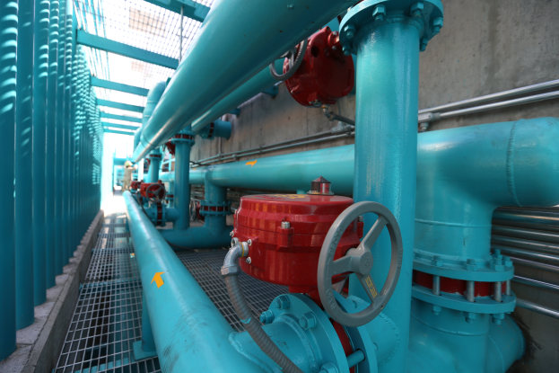 Eine große Gruppe von blauen Rohren und Ventilen in einem Gebäude mit einem prominenten roten Ventil in der Mitte, Metallstangen links und einer Wand im Hintergrund, die wahrscheinlich den Bau einer Gasleitung darstellt.