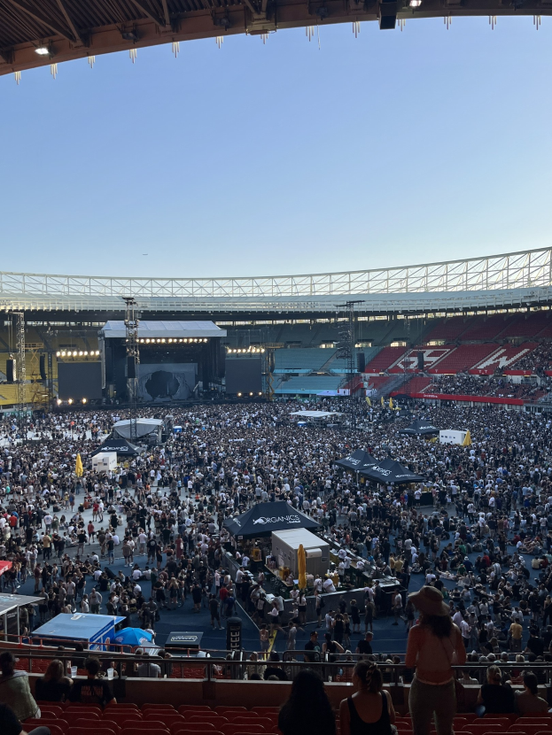Eine große Menschenmenge sitzt und steht in einem Stadion, umgeben von Geländern, Zelten und Lichtern, mit einem Dach und Himmel im Hintergrund.