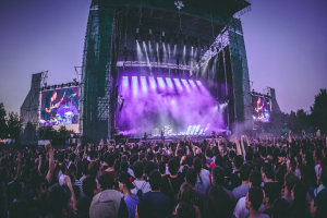 Eine große Menschenmenge versammelt sich auf einem Musikfestival, mit einer Bühne in der Mitte, auf der Musiker Instrumente spielen, beleuchtet von Scheinwerfern und umgeben von Bildschirmen, und einem Gebäude, Bäumen und einem klaren blauen Himmel im Hintergrund.