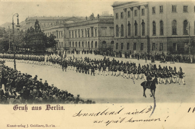 Ein Schwarz-Weiß-Foto einer Parade in Berlin, Deutschland, mit Menschen zu Fuß und zu Pferd, einer Statue, Straßenlaternen, Bäumen, Gebäuden und einem bewölkten Himmel, mit Text am unteren Rand.