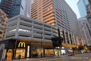 Ein McDonald's-Restaurant auf einer belebten Stadtstraße, flankiert von hohen Gebäuden, Straßenlaternen und -masten, sowie Fahrzeugen auf der Straße, Fußgängern auf dem Gehweg, Bäumen und einem klaren blauen Himmel.