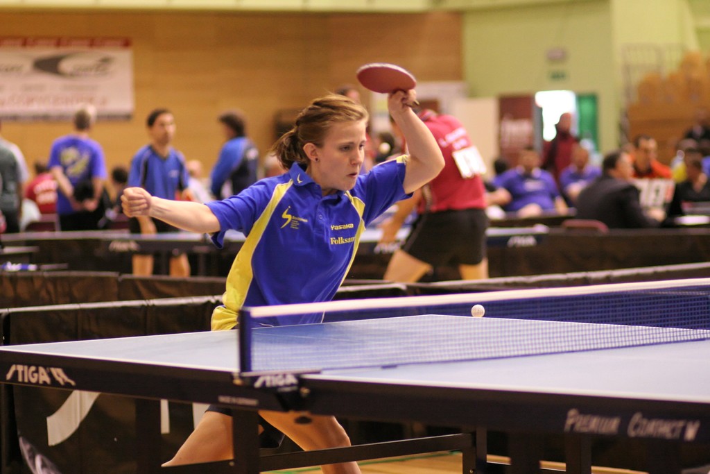 Eine Frau wird hervorgehoben, während sie einen Schläger hält und Tischtennis spielt, umgeben von ein paar sitzenden und stehenden Personen und einem Banner an der Wand.