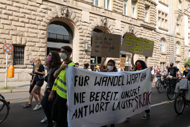 Eine Gruppe von Menschen marschiert auf der Straße in Berlin, hält Schilder und Banner hoch und fährt mit Fahrrädern vorbei an einem Gebäude mit Fenstern, Bögen, Säulen, Skulpturen und Bäumen.
