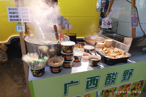 Ein Mann steht vor einem chinesischen Straßenfood-Wagen, bereitet mit verschiedenen Utensilien und Gegenständen Essen zu, mit Informationsschildern und einem Fahrzeug im Hintergrund.