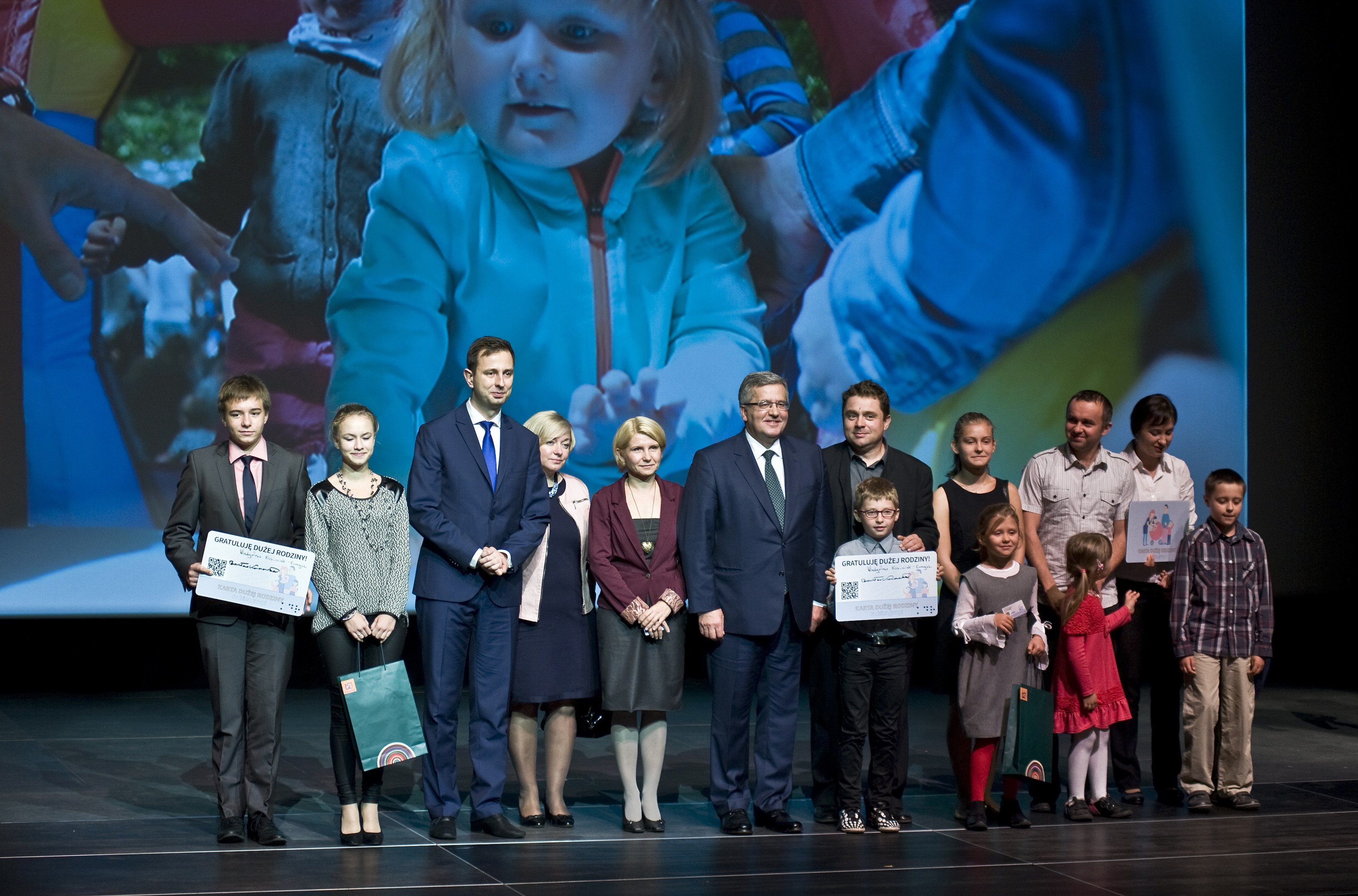 Eine Gruppe von Menschen auf einer Bühne mit Taschen und Urkunden, wobei ein Bildschirm ein Bild eines Kindes und andere Personen im Hintergrund anzeigt.