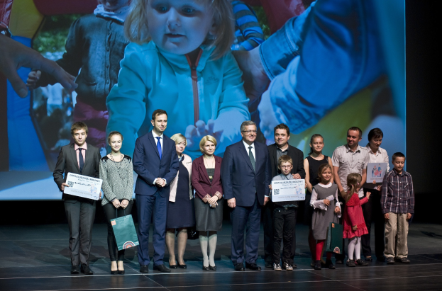 Eine Gruppe von Menschen auf einer Bühne mit Taschen und Urkunden, wobei ein Bildschirm ein Bild eines Kindes und andere Personen im Hintergrund anzeigt.