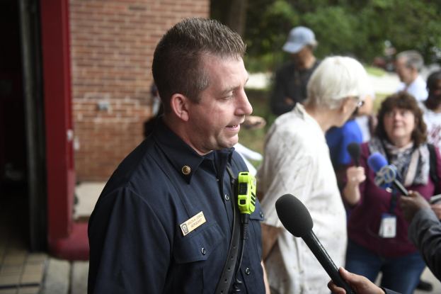Ein Polizist in Uniform spricht zu einer Gruppe von Menschen mit Mikrofonen, mit Bäumen und einem Gebäude im Hintergrund.