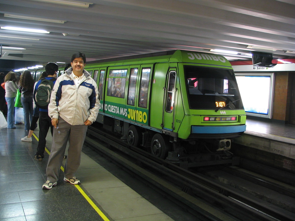 Ein Zug auf einem Bahngeleis mit Menschen auf der linken Seite und einem Dach oben im Bild.