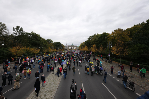 Eine große Gruppe von Menschen marschiert eine baumbestandene Straße in Berlin entlang, hält Kameras, mit einem Gebäude und einem klaren Himmel im Hintergrund.