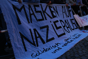 Eine Gruppe von Menschen, die auf dem Boden stehen und ein Banner mit der Aufschrift 'Masken auf Nazi-Partei' halten, vor einem Zaun mit Bäumen im Hintergrund.
