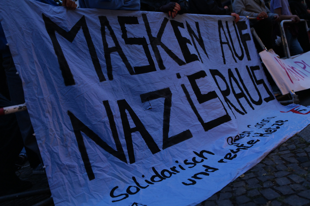 Eine Gruppe von Menschen, die auf dem Boden stehen und ein Banner mit der Aufschrift 'Masken auf Nazi-Partei' halten, vor einem Zaun mit Bäumen im Hintergrund.