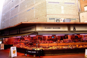 Ein belebter Markt mit verschiedenen Speisen, beleuchtet von Deckenlampen, umgeben von Texttafeln, vor Gebäuden unter einem klaren blauen Himmel.