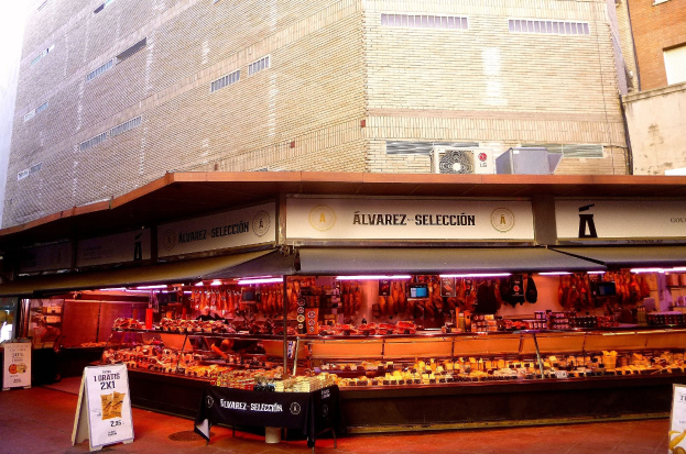 Ein belebter Markt mit verschiedenen Speisen, beleuchtet von Deckenlampen, umgeben von Texttafeln, vor Gebäuden unter einem klaren blauen Himmel.