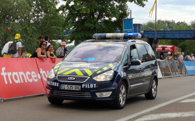 Polizeiauto fährt auf einer Straße neben einer Menschenmenge, mit einem Banner auf der linken Seite des Autos und Geländern mit Bannern dahinter, Bäumen, einer Brücke, einer Fahne und einem bewölkten Himmel im Hintergrund.