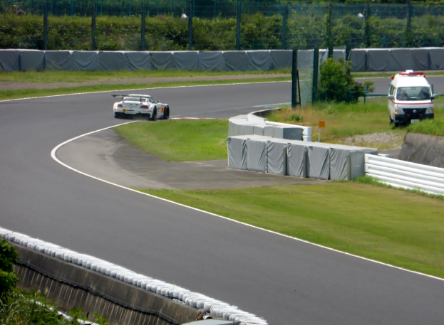 Ein Rennwagen fährt eine Rennstrecke entlang, im Hintergrund ist ein Polizeiwagen zu sehen, umgeben von Gras, Pflanzen, Bäumen und Absperrungen, mit einem Fahrzeug auf der rechten Seite und Text unten.