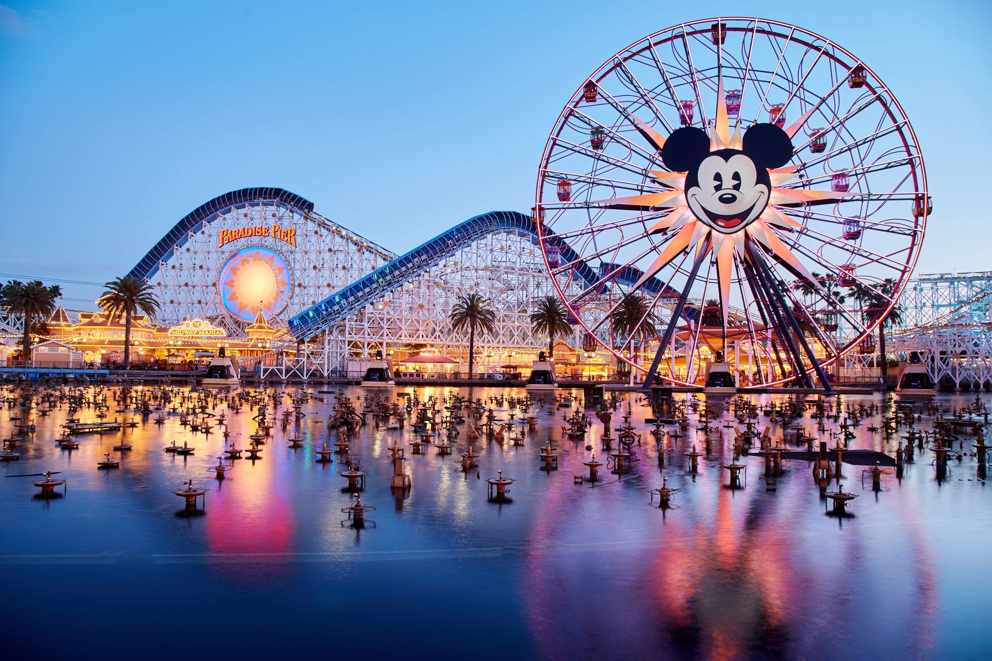 Eine lebendige Dämmerungsszene im Disneyland California Adventure mit einem riesigen Mickey-Maus-Rad im Vordergrund, einer Achterbahn im Hintergrund und Booten auf dem Wasser sowie einem Himmel aus Orangen, Pink- und Purpurtönen und silhouettenartigen Bäumen.