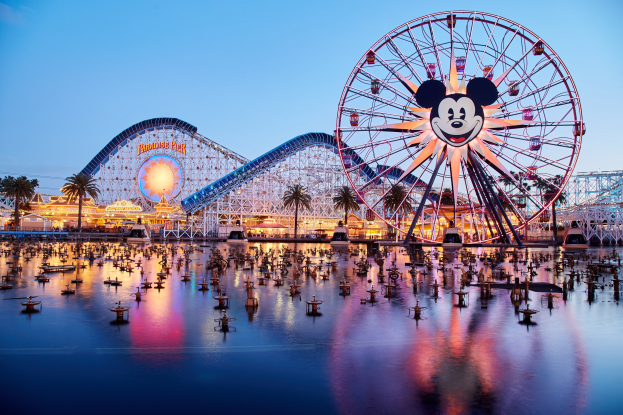 Eine lebendige Dämmerungsszene im Disneyland California Adventure mit einem riesigen Mickey-Maus-Rad im Vordergrund, einer Achterbahn im Hintergrund und Booten auf dem Wasser sowie einem Himmel aus Orangen, Pink- und Purpurtönen und silhouettenartigen Bäumen.