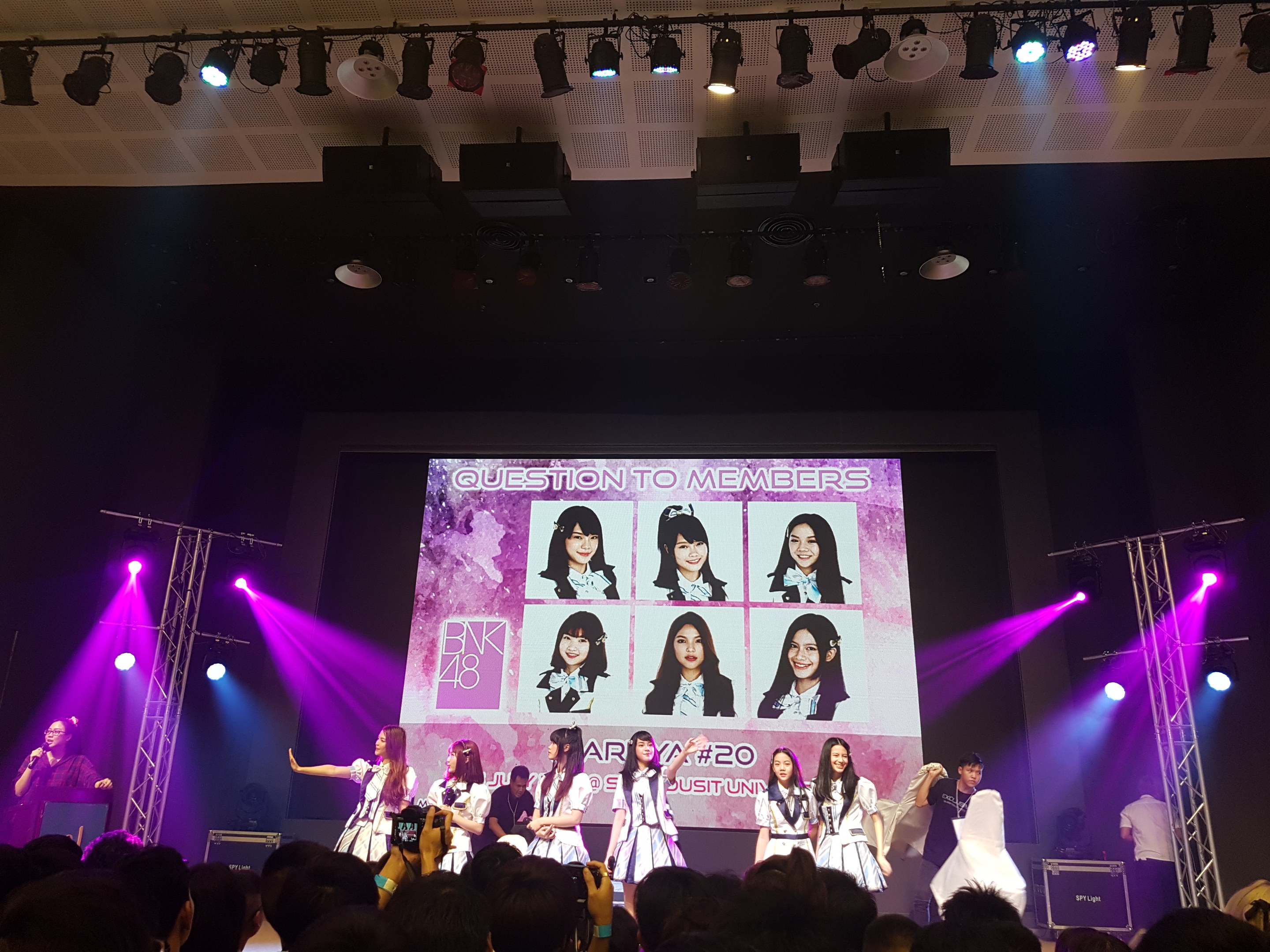 Eine Gruppe von Menschen, die auf einer hell erleuchteten Bühne performs, mit einer Menge darunter, mit einem Sänger mit Mikrofon und einem Musiker, der ein Instrument spielt, und einem großen Bildschirm im Hintergrund, der Bilder von BK48 anzeigt.