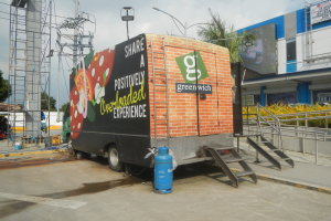 Lastwagen vor einem Gebäude mit einer Tafel, auf der "Teilen Sie eine positiv überladene Erfahrung" steht, mit Stufen, einem Zylinder am Boden, Geländern, Strommasten mit Drähten, Laternen, Bäumen und einer bewölkten Himmel.