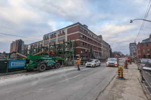 Eine Baustelle mit parkenden Autos, Verkehrszeichen, einer Person auf dem Gehweg, einem Kran, einem Zaun, einer Tafel, Pfosten, Drähte, Bäume, Gebäude und einem bewölkten Himmel.