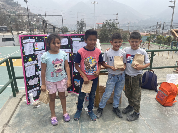 Gruppe von Kindern auf einem Basketballfeld mit Papiertüten in den Händen, Tüten und Gegenstände rechts und eine Tafel mit Text links, vor dem Hintergrund von Zäunen, Pfosten, Laternen, Bäumen, Gebäuden, Hügeln und Himmel.