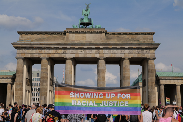 Eine Gruppe von Menschen steht vor dem Brandenburger Tor in Berlin, Deutschland, mit einer "Racial Justice"-Schriftzug-Halterung, wobei die Säulen und die Statue des Tors sowie Gebäude und ein bewölkter Himmel im Hintergrund zu sehen sind.