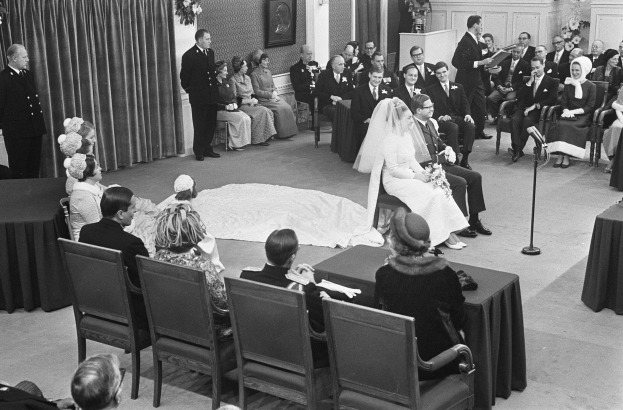 Schwarze und weiße Hochzeitszeremonie mit sitzenden und stehenden Menschen, ein Mann spricht in ein Mikrofon, eine Frau hält einen Blumenstrauß, ein Podium mit einem Mikrofon, Blumengestecke, ein Vorhang, ein gerahmtes Foto an der Wand und eine Tür im Hintergrund.