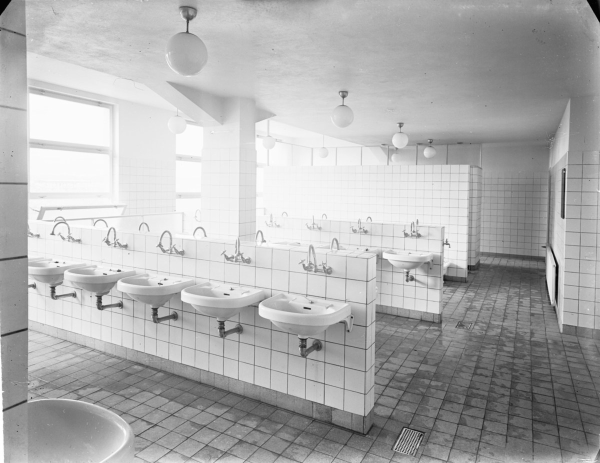 Schwarzes und weißes Foto einer öffentlichen Toilette mit Waschbecken, Armaturen, Wänden, Beleuchtung und Fenstern.