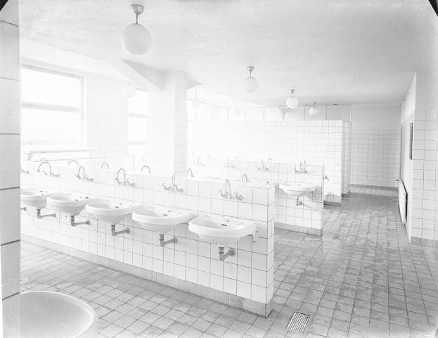 Schwarzes und weißes Foto einer öffentlichen Toilette mit Waschbecken, Armaturen, Wänden, Beleuchtung und Fenstern.