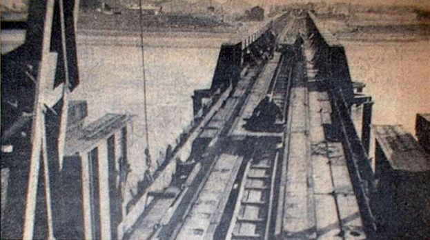 Ein Schwarz-Weiß-Foto einer Bahntrasse mit einer im Hintergrund befindlichen Brücke, umgeben von verstreuten Werkzeugen und Materialien.
