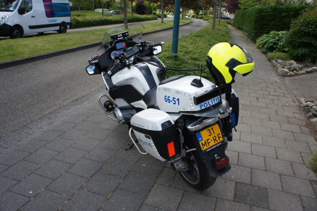 Eine Polizeimotorrad auf dem Seitenstreifen geparkt, mit einem Helm darauf, umgeben von Bäumen, Pfählen, Gebäuden, Pflanzen, Gras, Steinen und anderen Fahrzeugen im Hintergrund.