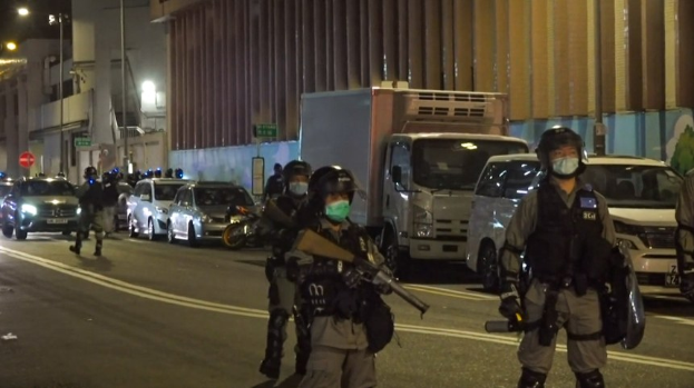 Gruppe von Polizisten in Helmen, Handschuhen und Gesichtsmasken, die auf einer Straße in der Nacht stehen, einige halten Waffen, mit Fahrzeugen, Laternen, Schildern und Gebäuden im Hintergrund.