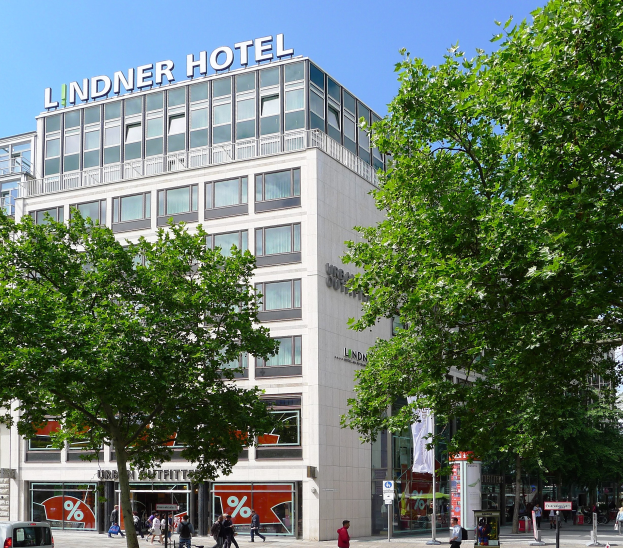Das Lindner Hotel in Berlin, ein großes Glasfrontgebäude mit Aufschrift auf der Oberseite, umgeben von Bäumen, Fahrzeugen und Passanten unter einem sichtbaren Himmel.